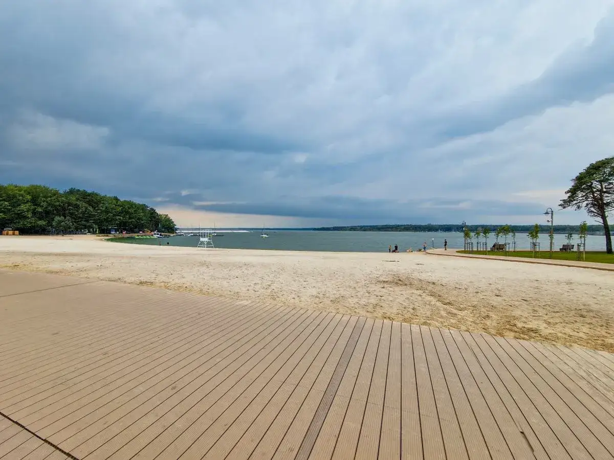 Plaża nad zalewem sulejowskim z drewnianym molo. W oddali widać wodę, łodzie i las.