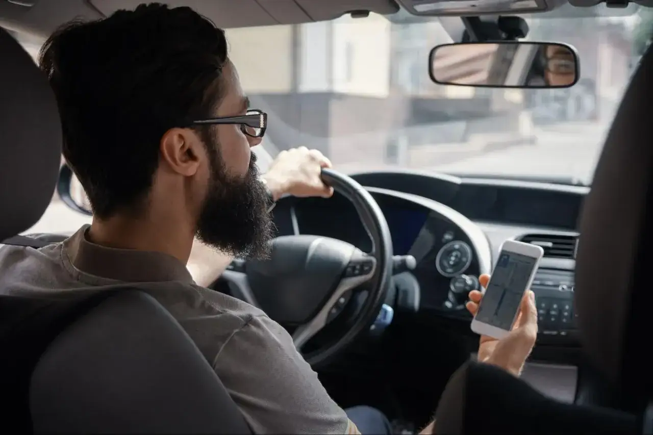 Mężczyzna z brodą i okularami prowadzi samoch&oacute;d, trzymając telefon z nawigacją. Sprawdza trasę, by zminimalizować prowizję Uber.