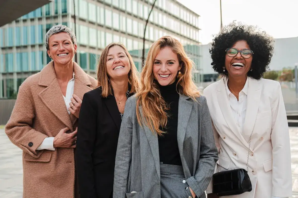 Vier Frauen in Business-Casual-Kleidung lachen vor einem modernen Geb&auml;ude.