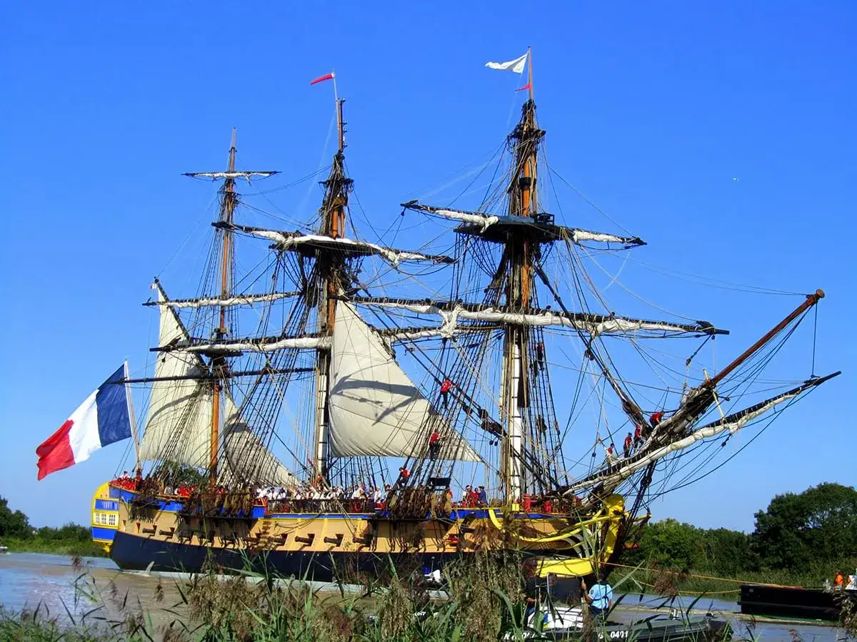 Le majestueux bateau l'Hermione, réplique d'une frégate du XVIIIe siècle, navigue fièrement sous un ciel bleu, pavillon français au vent.