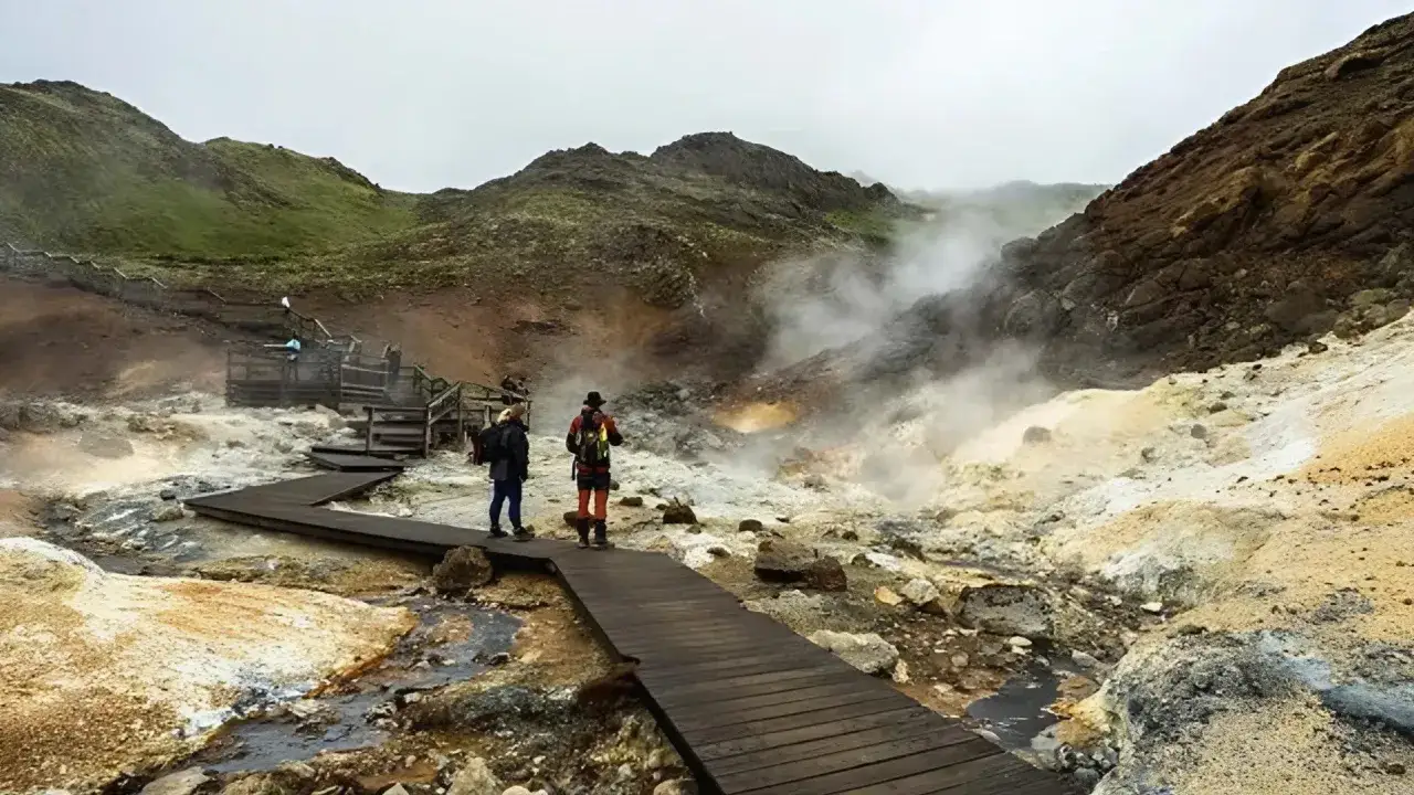 Para spacerowa prowadzi przez parujące, geotermalne tereny. W oddali widać wzgórza, a w powietrzu unosi się mgiełka, przypominająca tę z **blue lagoon**.