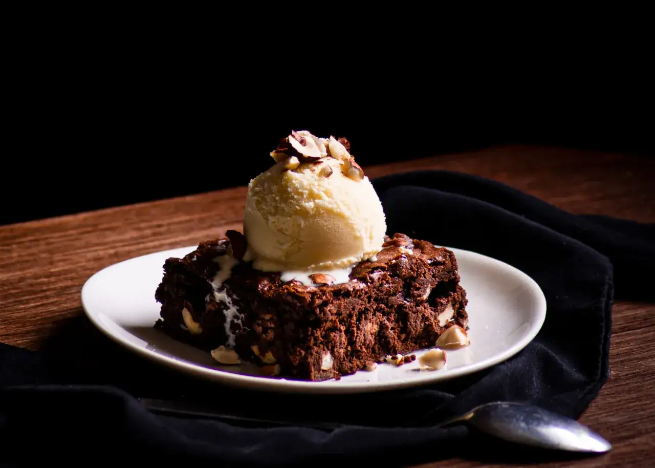 Un delicioso brownie de chocolate con una bola de helado de vainilla y nueces picadas, servido en un plato blanco.