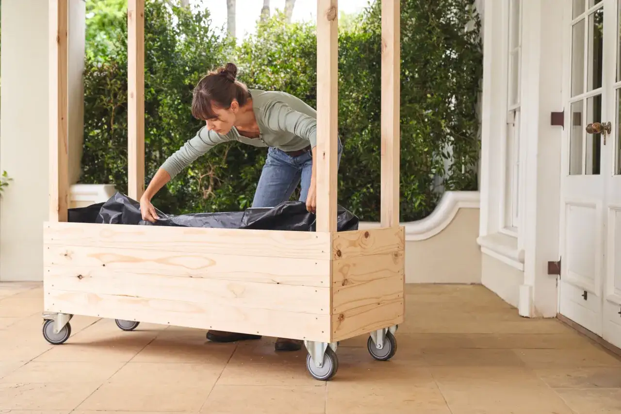 Une femme installe une bâche noire dans une jardinière en bois à roulettes, étape clé pour comment fabriquer toit pour tomates.