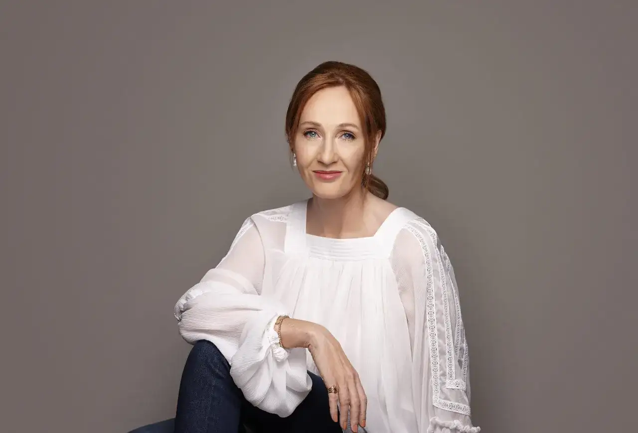 J.K. Rowling, autorka Harrego Pottera, w białej bluzce i dżinsach, z uśmiechem patrzy w obiektyw.