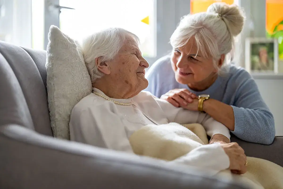 Una donna anziana, forse affetta da demenza senile, sorride mentre un'altra donna le tiene la mano con affetto.