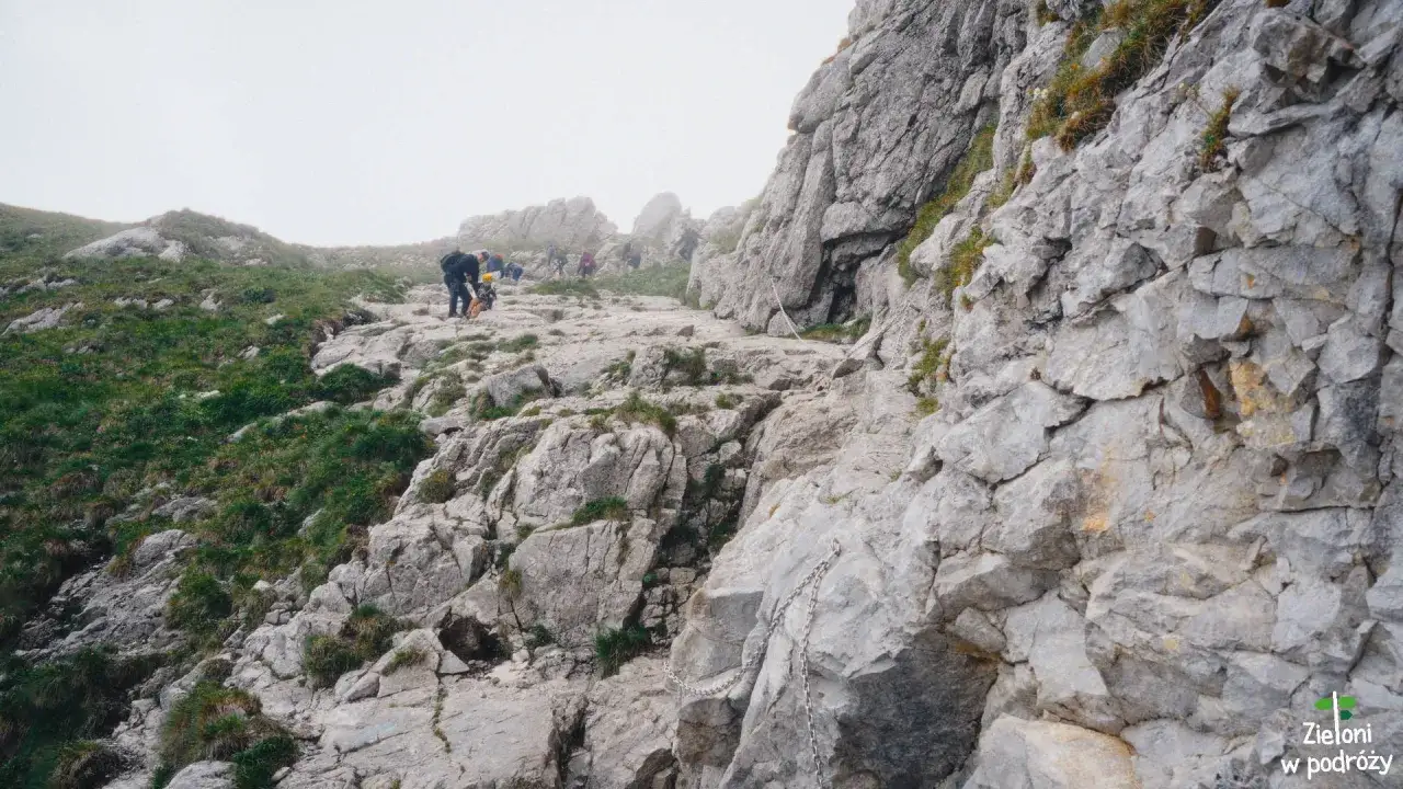 Grupa turyst&oacute;w wspina się po kamienistym szlaku. W oddali majaczy mglisty **wielki Giewont**.