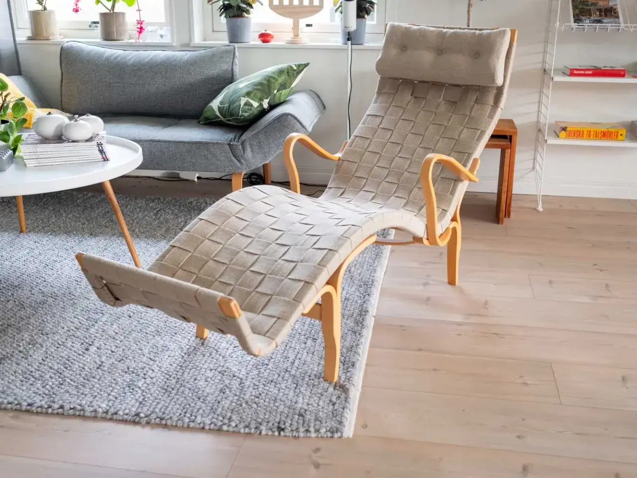 En klassisk bruno mathsson liggf&aring;t&ouml;lj i beige fl&auml;tat tyg med b&ouml;jda tr&auml;armst&ouml;d, placerad p&aring; en gr&aring; matta i ett ljust vardagsrum.