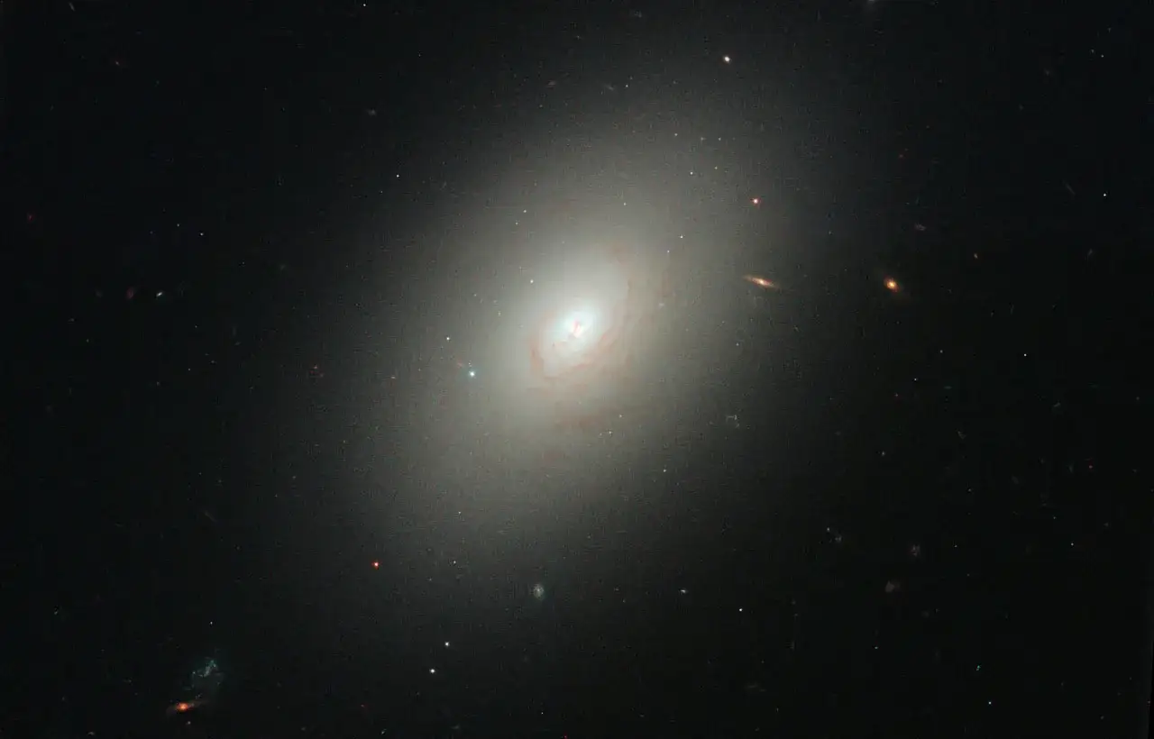 En stor, ljus elliptisk galax dominerar bilden, omgiven av mindre galaxer och stj&auml;rnor.