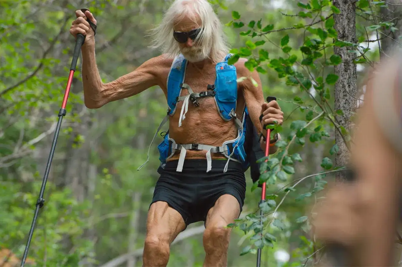 Un uomo anziano con capelli lunghi e bianchi, occhiali da sole e barba, affronta un sentiero boscoso con bastoncini da trekking. Indossa uno zaino da corsa e pantaloncini neri, pronto per l'ultramaratona più lunga del mondo.