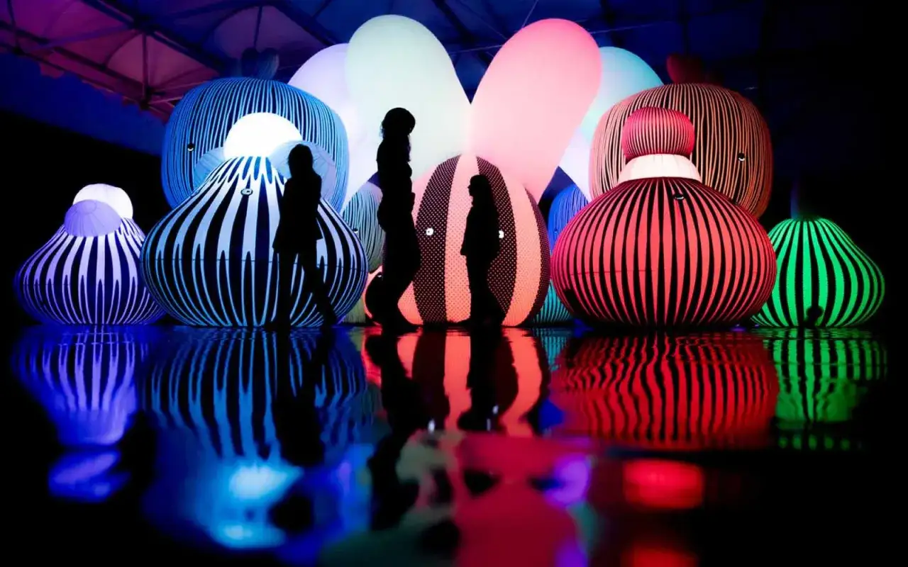 Silhouettes of people wander through a vibrant balloon museum in London, surrounded by glowing, striped inflatable sculptures.