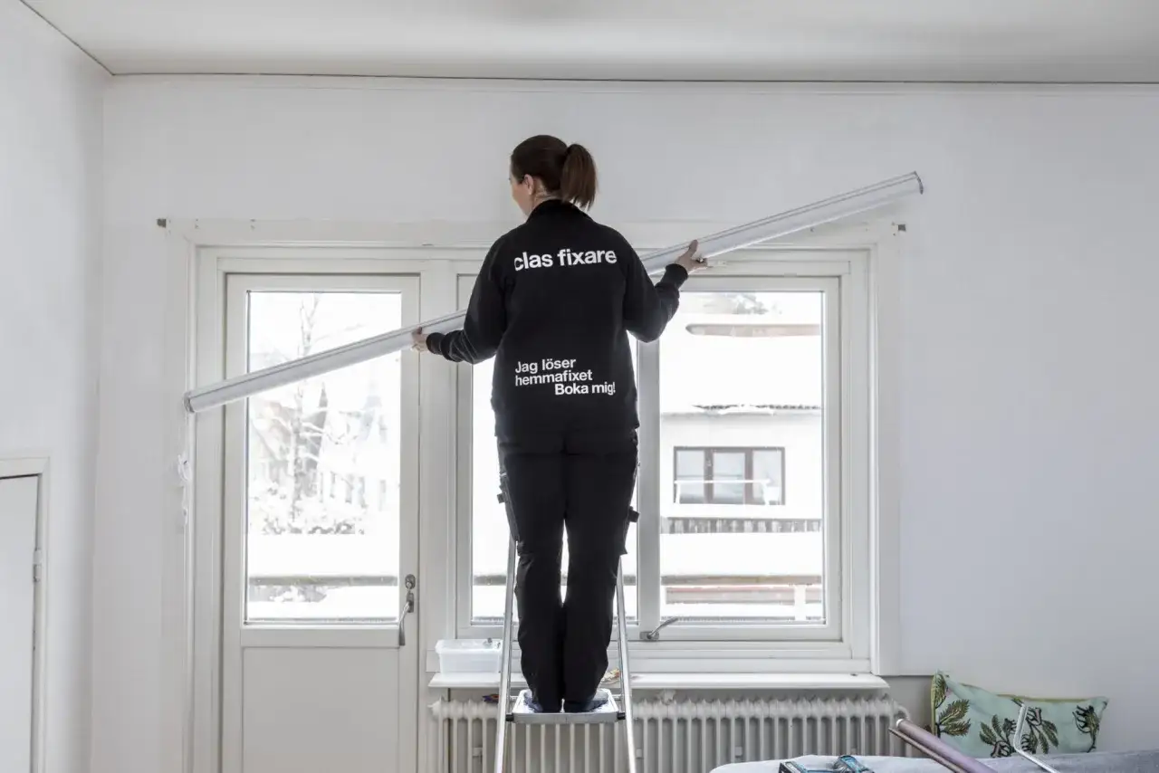En person på en stege monterar en gardinstång högt upp vid ett fönster.