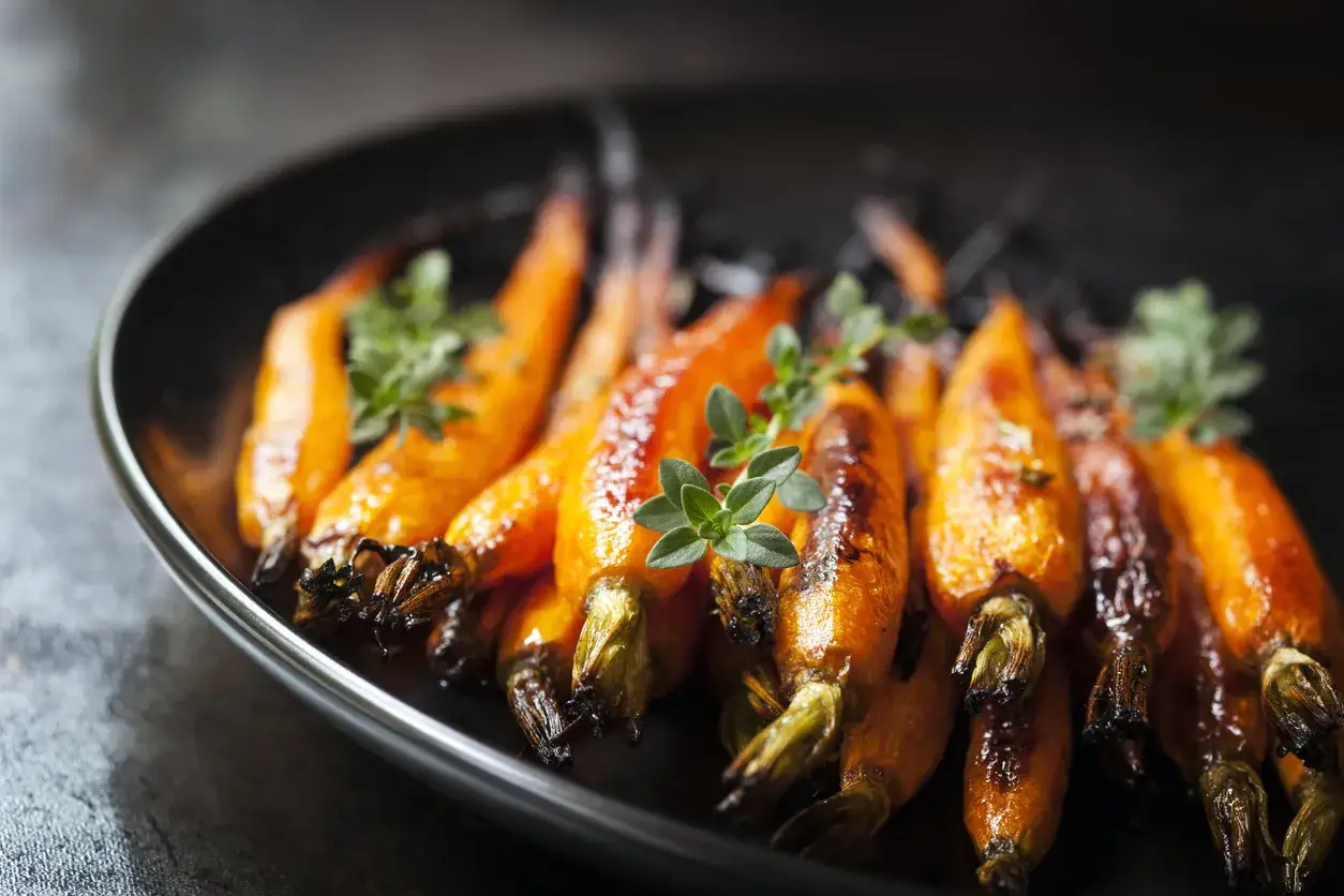 Carote caramellate e dorate, cotte in friggitrice ad aria, guarnite con erbe aromatiche fresche su un piatto scuro.