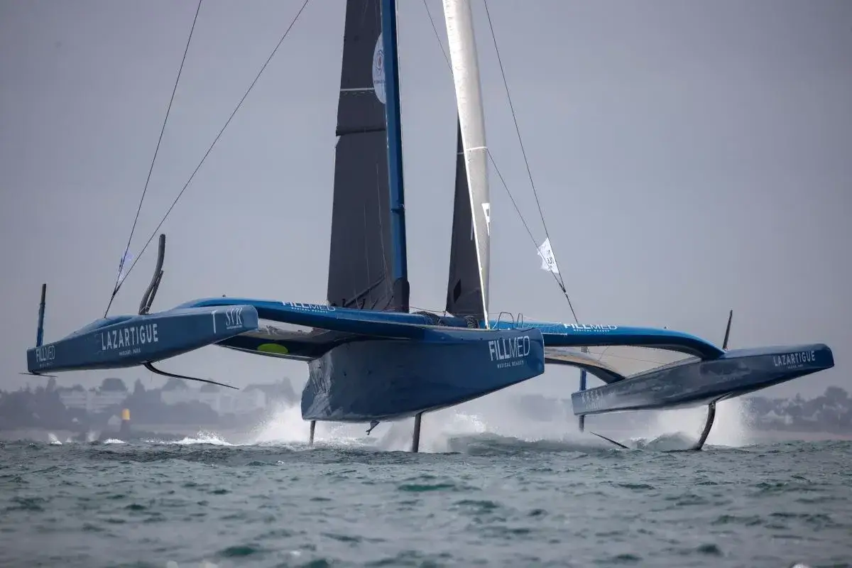 Le trimaran Gitana 18 fend les vagues, ses foils soulevant la coque. La mer est agitée, le ciel gris.