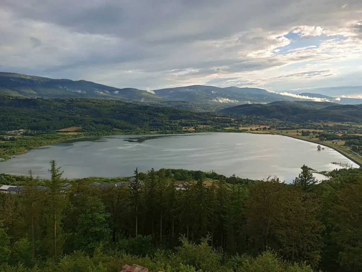 Rozległy zbiornik Sosnówka otoczony zielonymi wzgórzami i górami. Spokojna tafla wody odbija chmury na niebie.