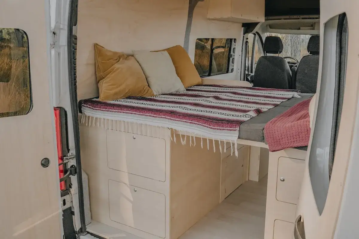 Aménagement intérieur d'un van avec un lit confortable, des coussins douillets et des rangements pratiques. Ce lit 1 place est parfait pour vos aventures.