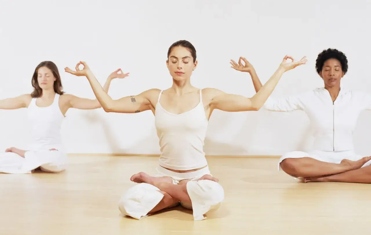 Drei Frauen in wei&szlig;er Kleidung praktizieren Kundalini Yoga im Lotussitz, die Augen geschlossen, die H&auml;nde in Mudras erhoben.