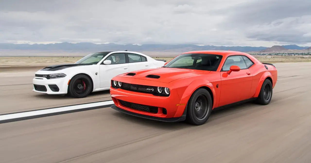 Dwa potężne muscle cary, pomarańczowy Dodge Challenger SRT i biały Charger SRT, pędzą po pustynnej drodze.