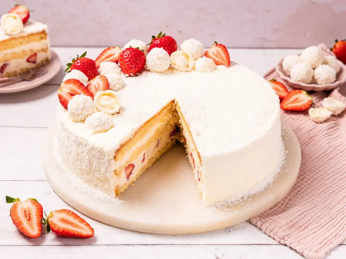 Eine köstliche Erdbeer Raffaello Torte, mit frischen Erdbeeren und Kokosraspeln verziert. Ein Stück ist bereits angeschnitten.