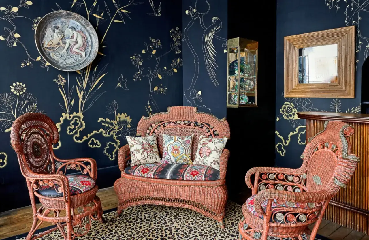 Un salon aux murs bleu nuit orn&eacute;s de motifs dor&eacute;s, avec du mobilier napol&eacute;on 3 en rotin corail, coussins color&eacute;s et tapis l&eacute;opard.