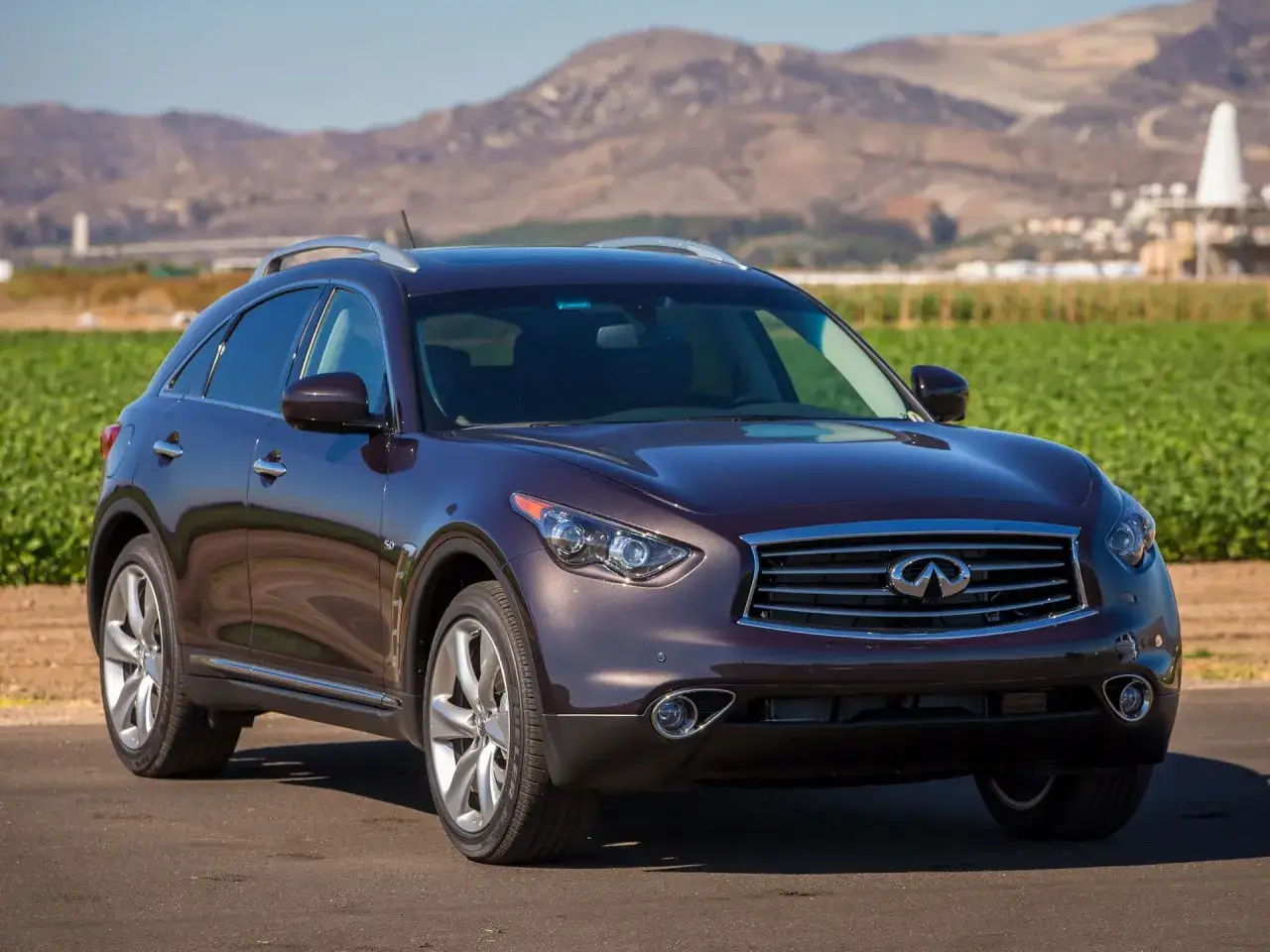 Elegancki, ciemnobordowy Infiniti QX70 na tle zielonych pól i gór.