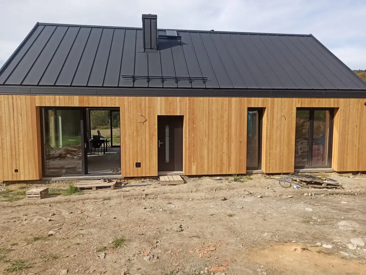 Elewacja z modrzewia syberyjskiego po latach, z ciemnym dachem i dużymi oknami. Wokół plac budowy.