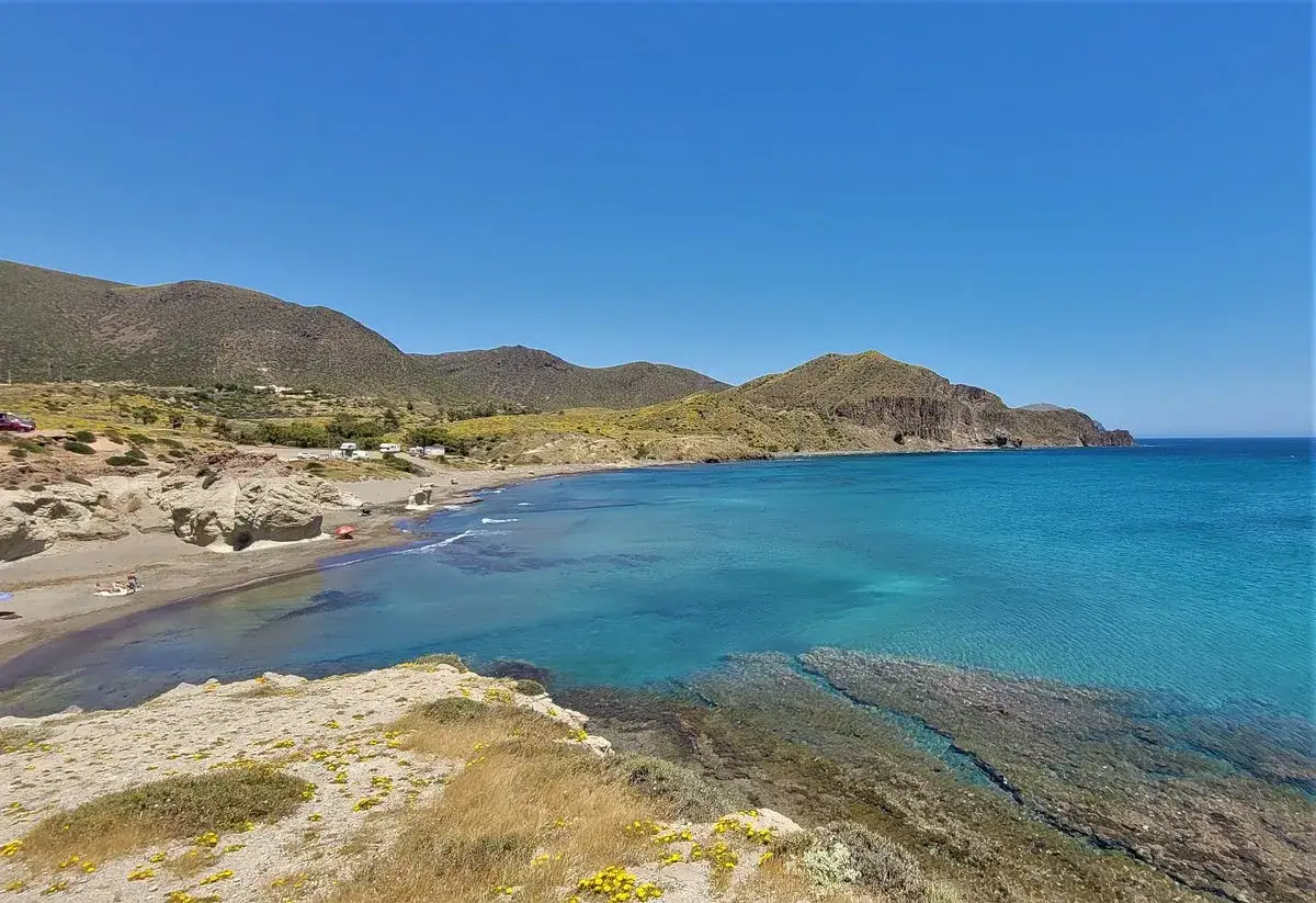 Aguas turquesas bañan la playa de **Isleta del Moro, Almería**, con montañas verdes y rocosas de fondo.