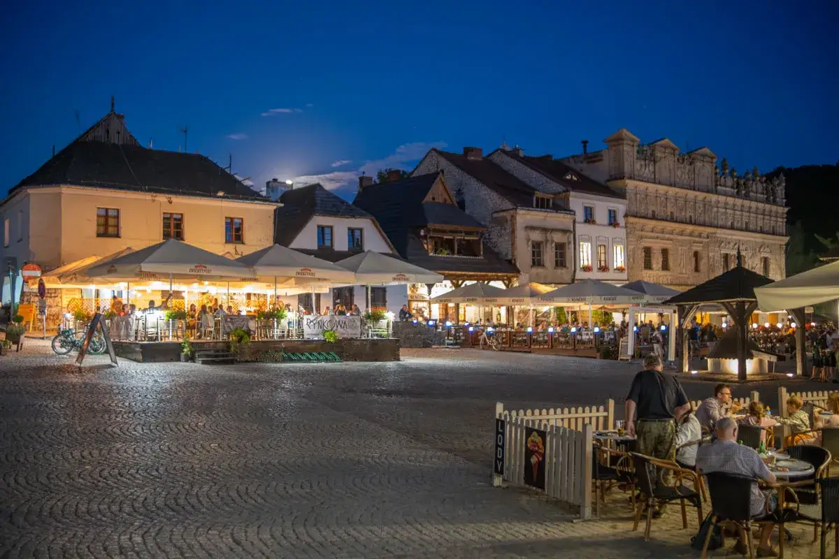 Wieczorny rynek Kazimierza Dolnego tętni życiem. Ludzie siedzą przy stolikach pod parasolami, ciesząc się atmosferą.