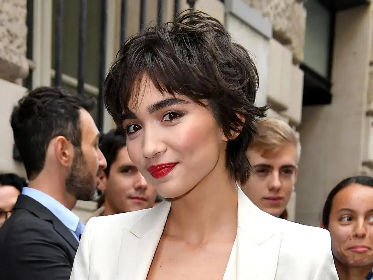 Eine Frau mit einer stylischen Frisur zwischen Pixie und Bob, rotem Lippenstift und einem wei&szlig;en Blazer.