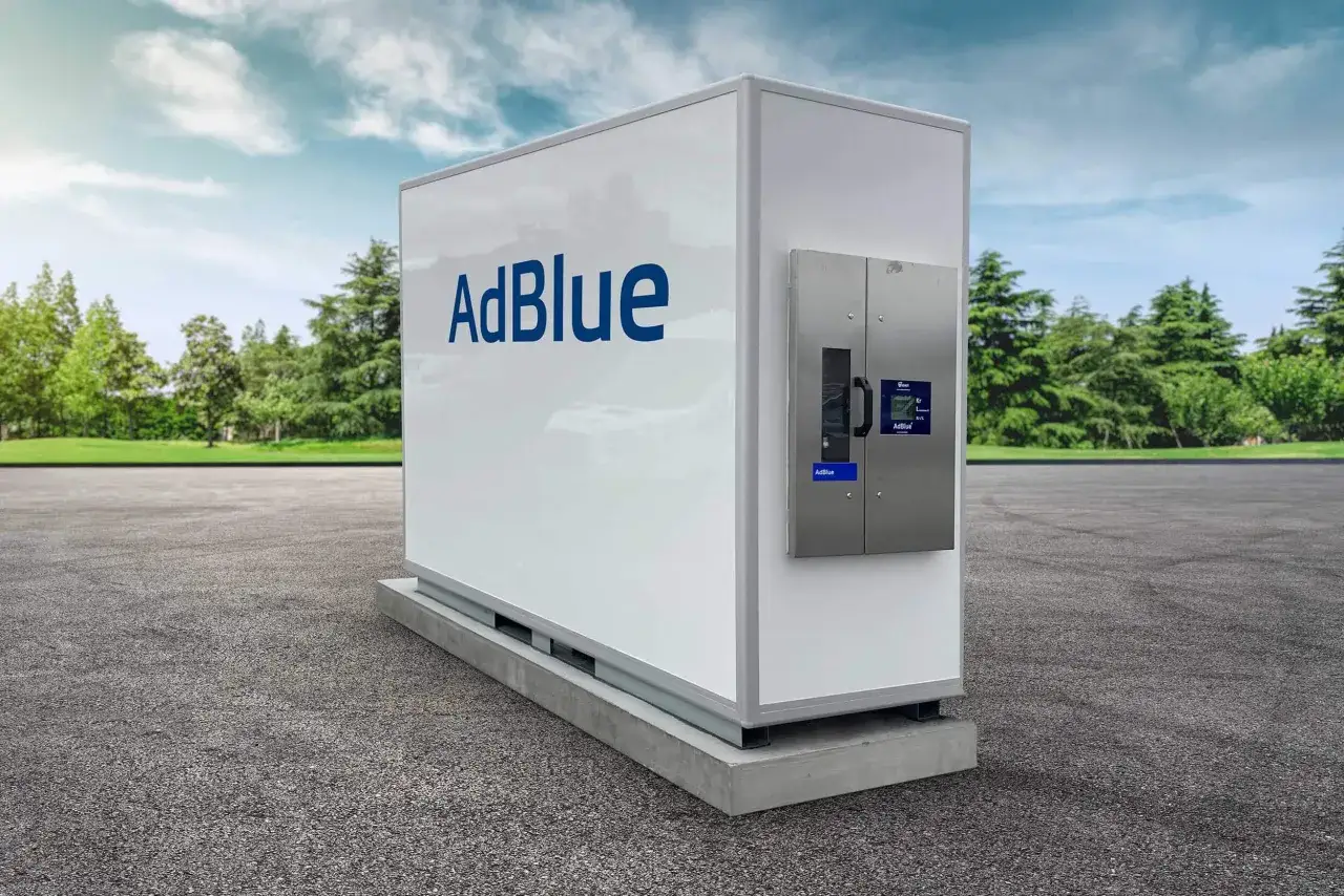 En vit adblue tankstation st&aring;r p&aring; en grusplan, omgiven av gr&ouml;na tr&auml;d och en bl&aring; himmel.