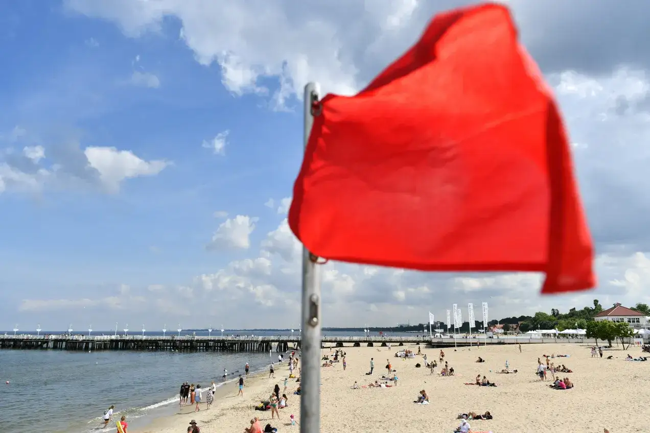 Czerwona flaga na plaży. Czy można się kąpać w Bałtyku? Dziś nie, bo woda jest wzburzona. Ludzie opalają się i spacerują.