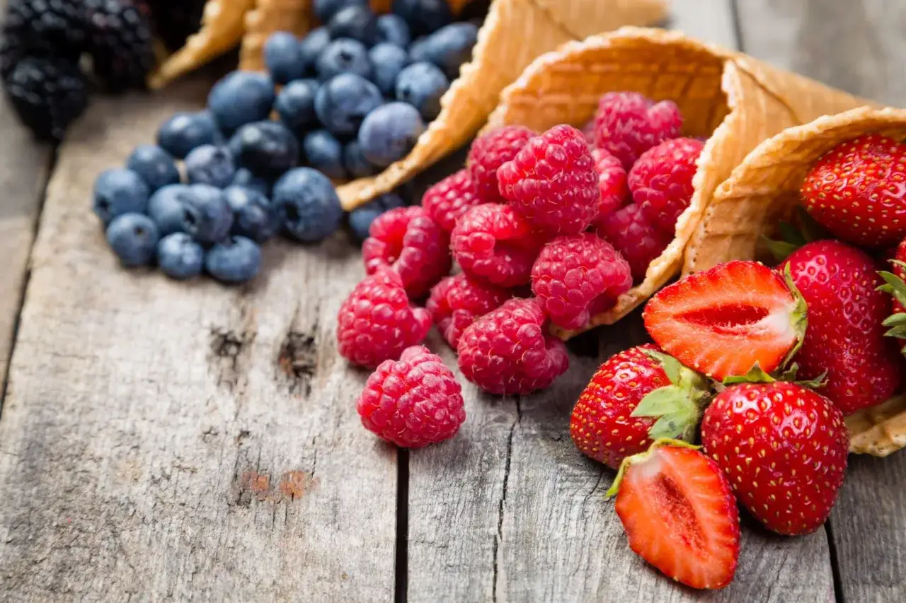 Sommerliche Beeren wie Himbeeren, Blaubeeren und Erdbeeren in Waffelhörnchen auf Holzuntergrund.