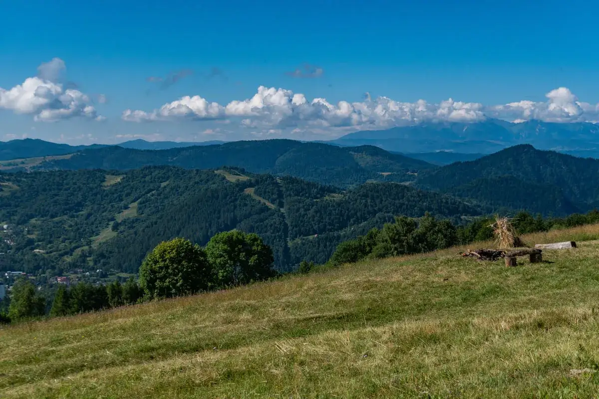 Widok na górzyste tereny Beskidu Sądeckiego z widoczną z daleka bacówką pod Bereśnikiem. Na pierwszym planie trawiasty stok, w tle lasy i niebieskie niebo z chmurami.