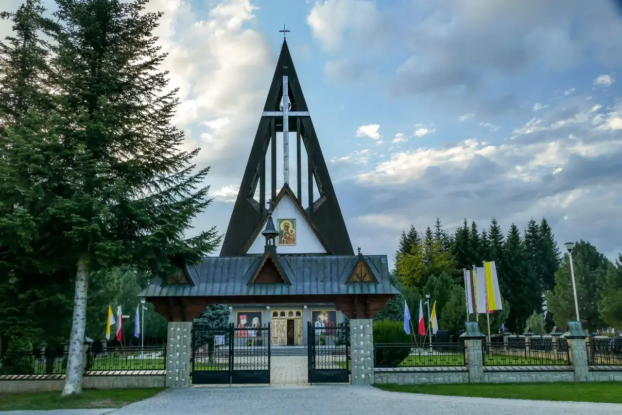 Nowoczesny kościół na Bachledówce z charakterystyczną, strzelistą wieżą i krzyżem. Przed świątynią brama i ogrodzenie.