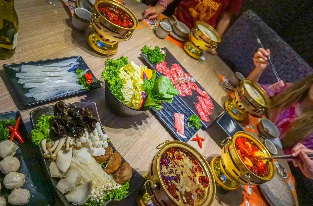Ein Tisch voller K&ouml;stlichkeiten: Was ist Hot Pot? Hier brutzeln Br&uuml;hen in goldenen T&ouml;pfen, umgeben von frischem Gem&uuml;se, d&uuml;nn geschnittenem Fleisch und Pilzen.