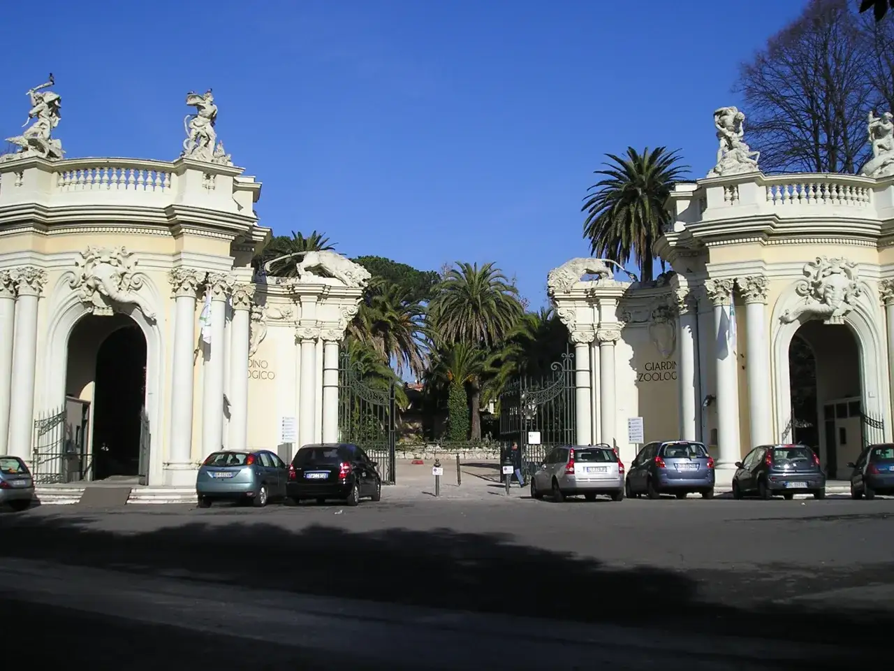 Monumentalne wejście do zoo w Rzymie z rzeźbami lwów i palmami.