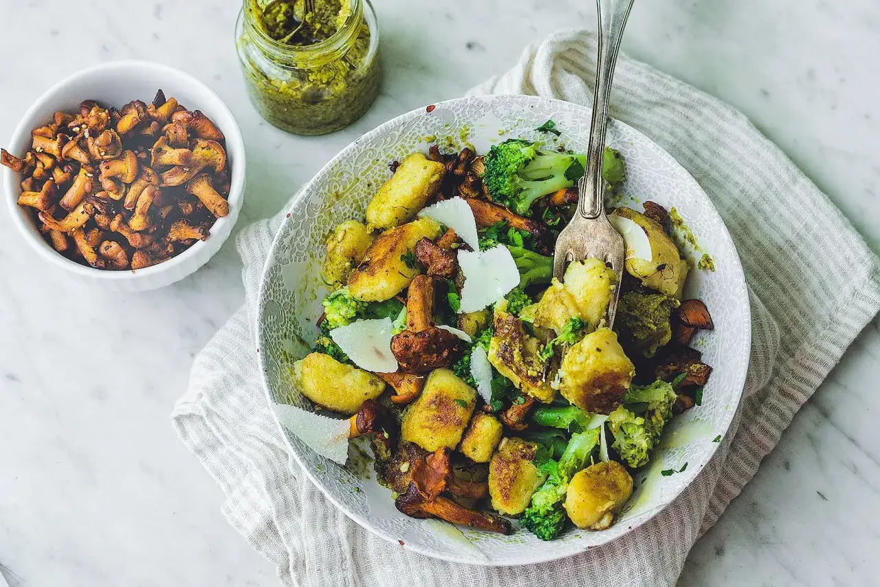 Pyszne gnocchi z kurkami, brokułami i parmezanem, polane zielonym pesto. Obok miseczka z kurkami i słoiczek pesto.