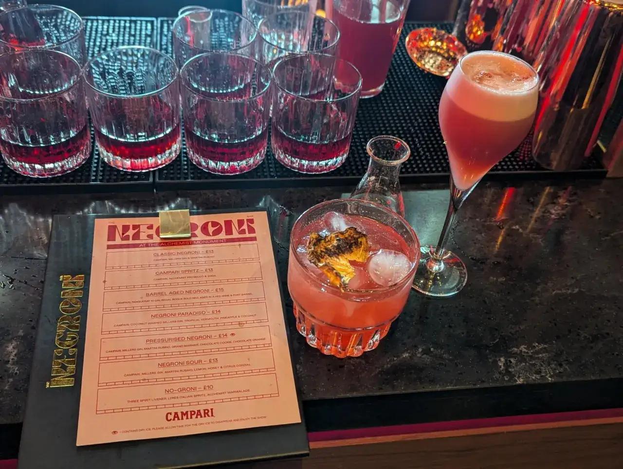 Menu z drinkami, gdzie widać kilka kieliszków z czerwonymi napojami i dwa koktajle. Na pierwszym planie menu z napisem