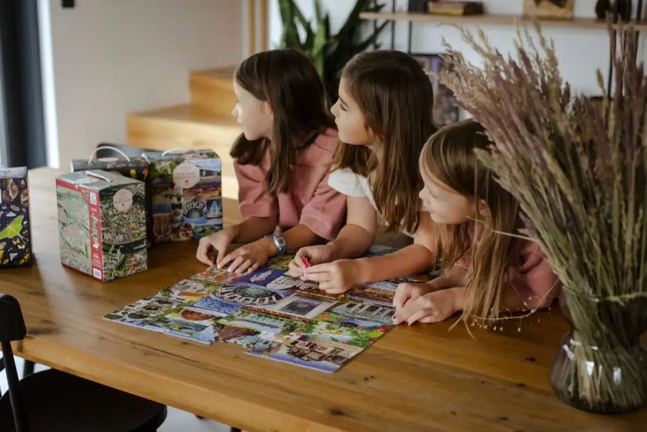 Trzy dziewczynki układają puzzle z widokami polskich miast. W tle pudełka z kolejnymi zestawami do układania.