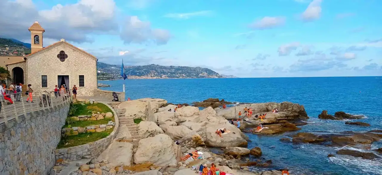 Chiesa e scogli a picco sul mare, gente che prende il sole. Un assaggio di cosa vedere a Bordighera, tra storia e relax balneare.