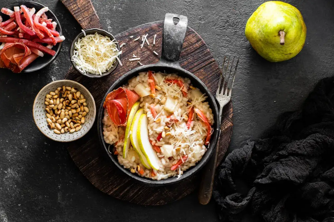 Risotto pere e speck cremoso, guarnito con fette di pera, speck croccante e pinoli tostati. Un piatto autunnale invitante.