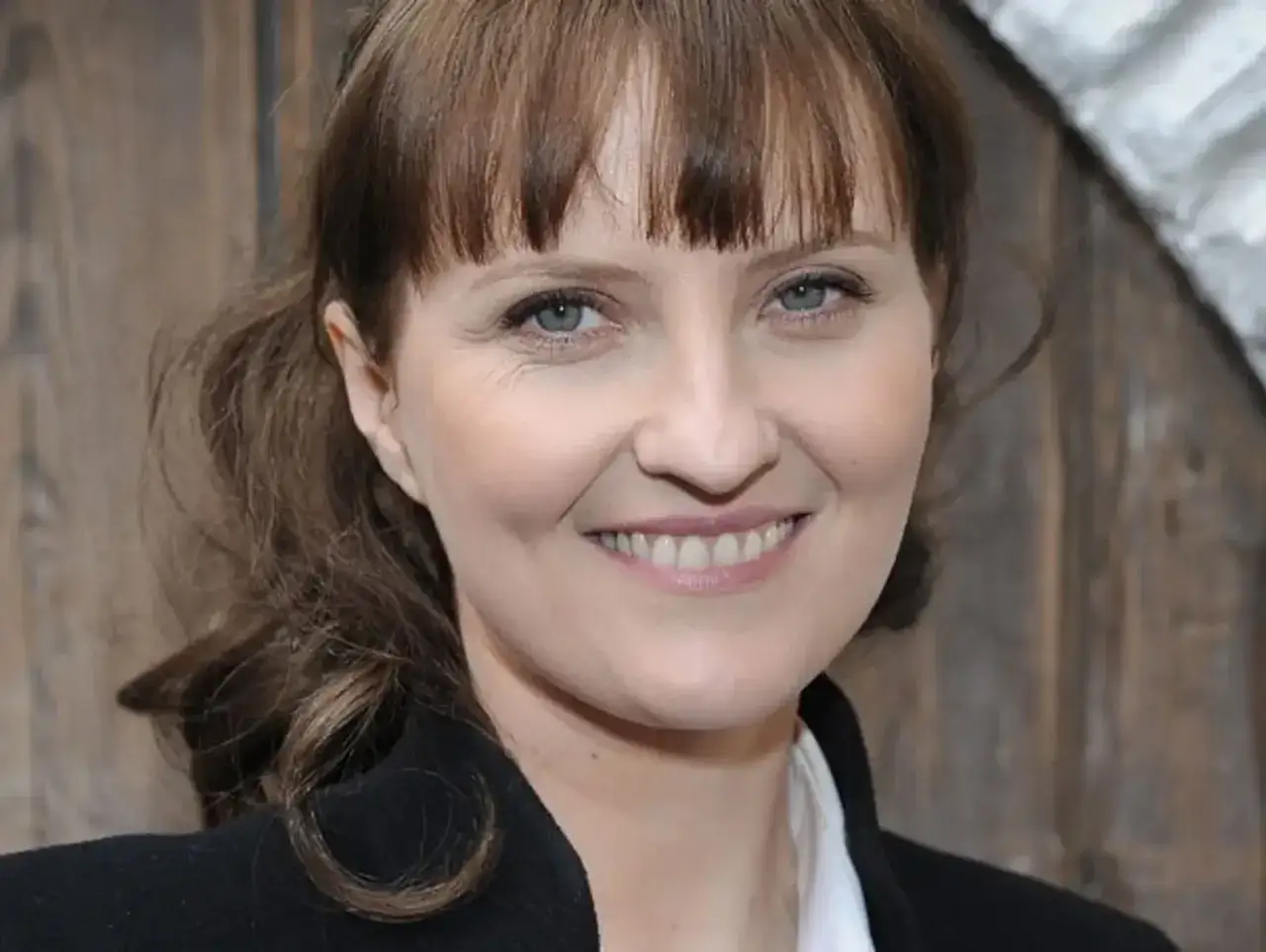 Uśmiechnięta Izabela Kuna młoda, z burzą brązowych włosów i niebieskimi oczami, w czarnym płaszczu.