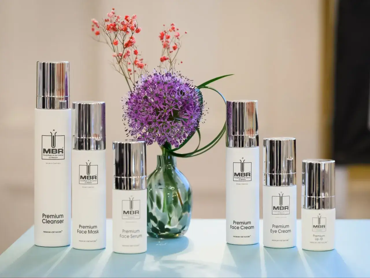 Linia kosmetyków MBR: Premium Cleanser, Face Mask, Face Serum, Face Cream, Eye Cream i Lip-ID, obok bukietu fioletowych i różowych kwiatów.