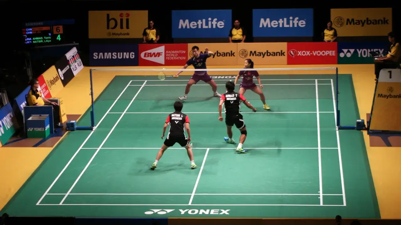 Mecz badmintonowy, gracze w akcji. Widoczni zawodnicy z Indonezji i Chin. W tle reklamy, w tym 