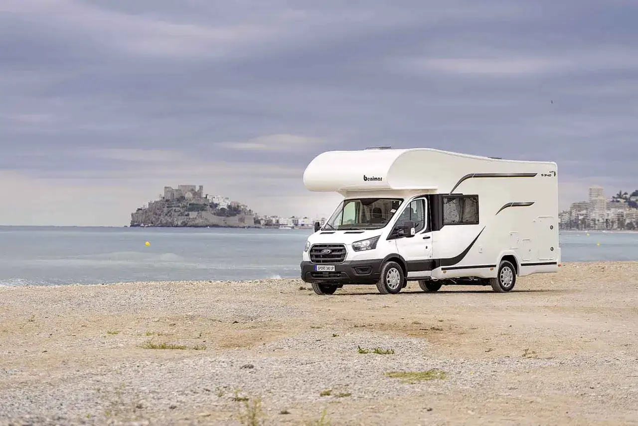 Camping-car Benimar blanc sur une plage de sable, avec un château en ruines et une ville côtière en arrière-plan.