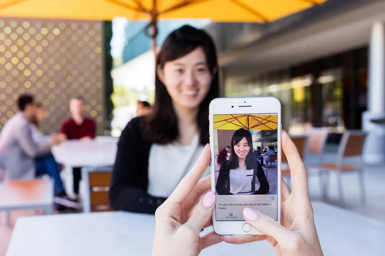 Una donna sorride mentre un telefono mostra la sua foto, con una didascalia generata da seeing AI.
