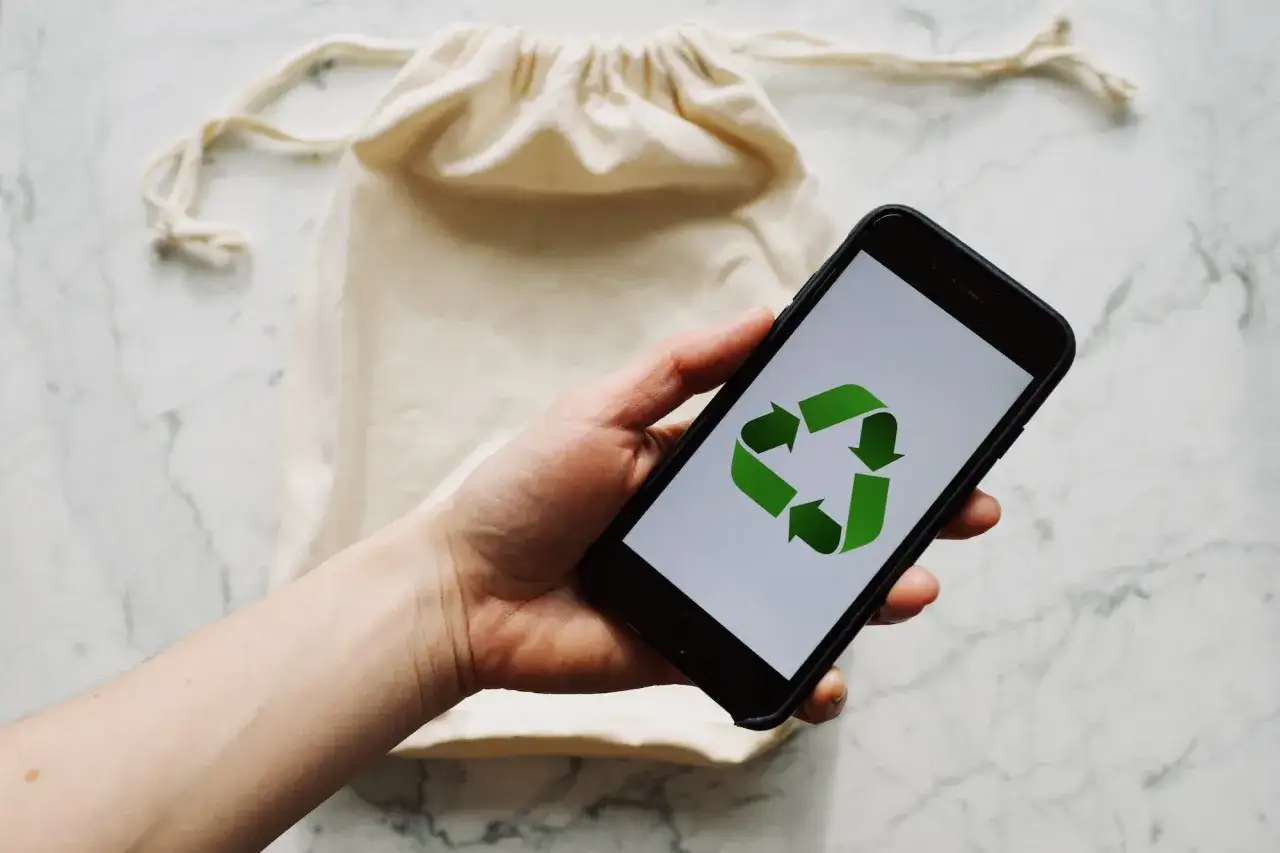 Dłoń trzyma smartfon z zielonym symbolem recyklingu treści. W tle beżowa torba.