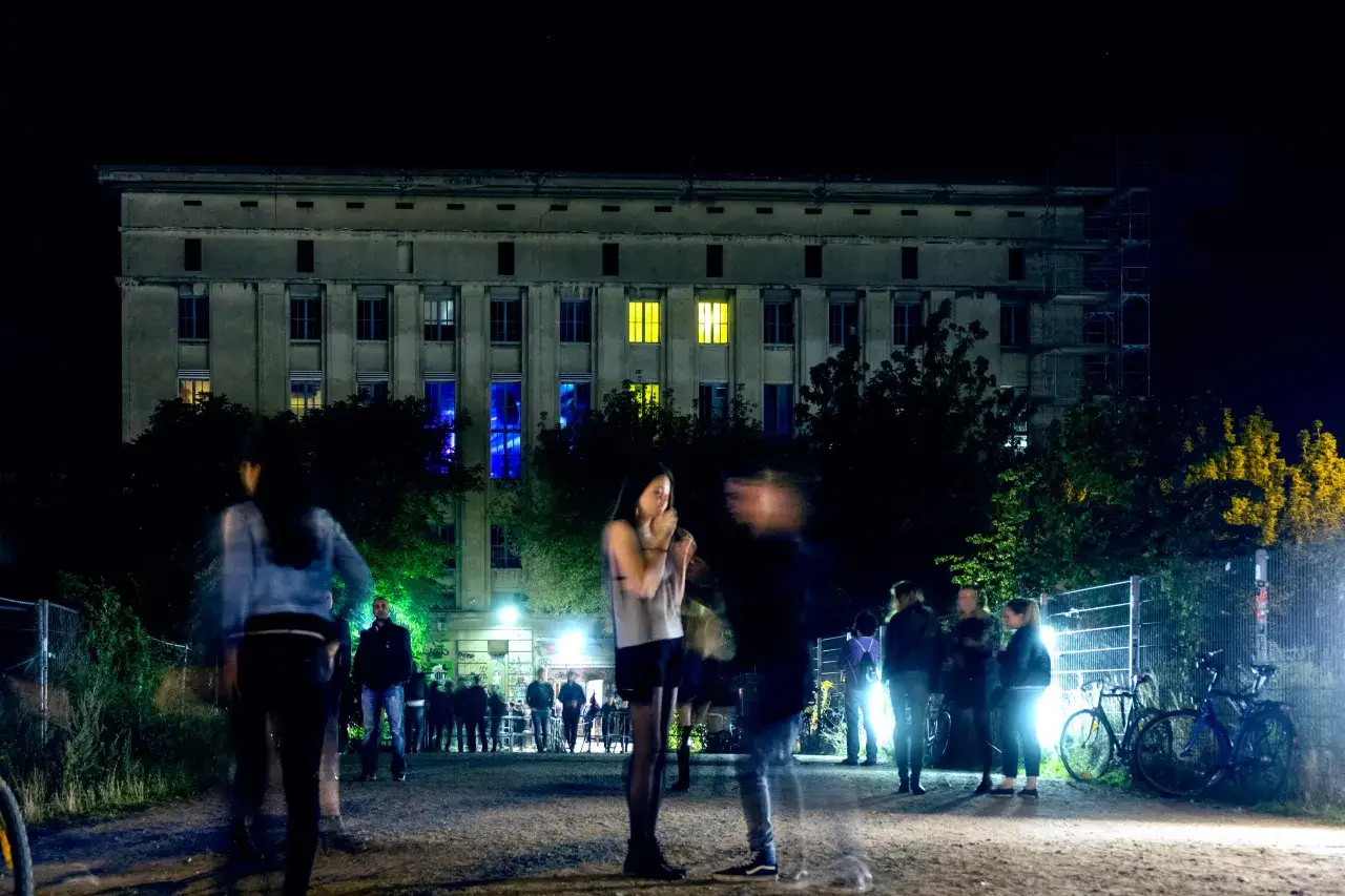 Persone sfocate davanti al Berghain di notte, con luci blu e gialle che filtrano dalle finestre. Un'atmosfera misteriosa, quasi un invito a capire come entrare al Berghain.
