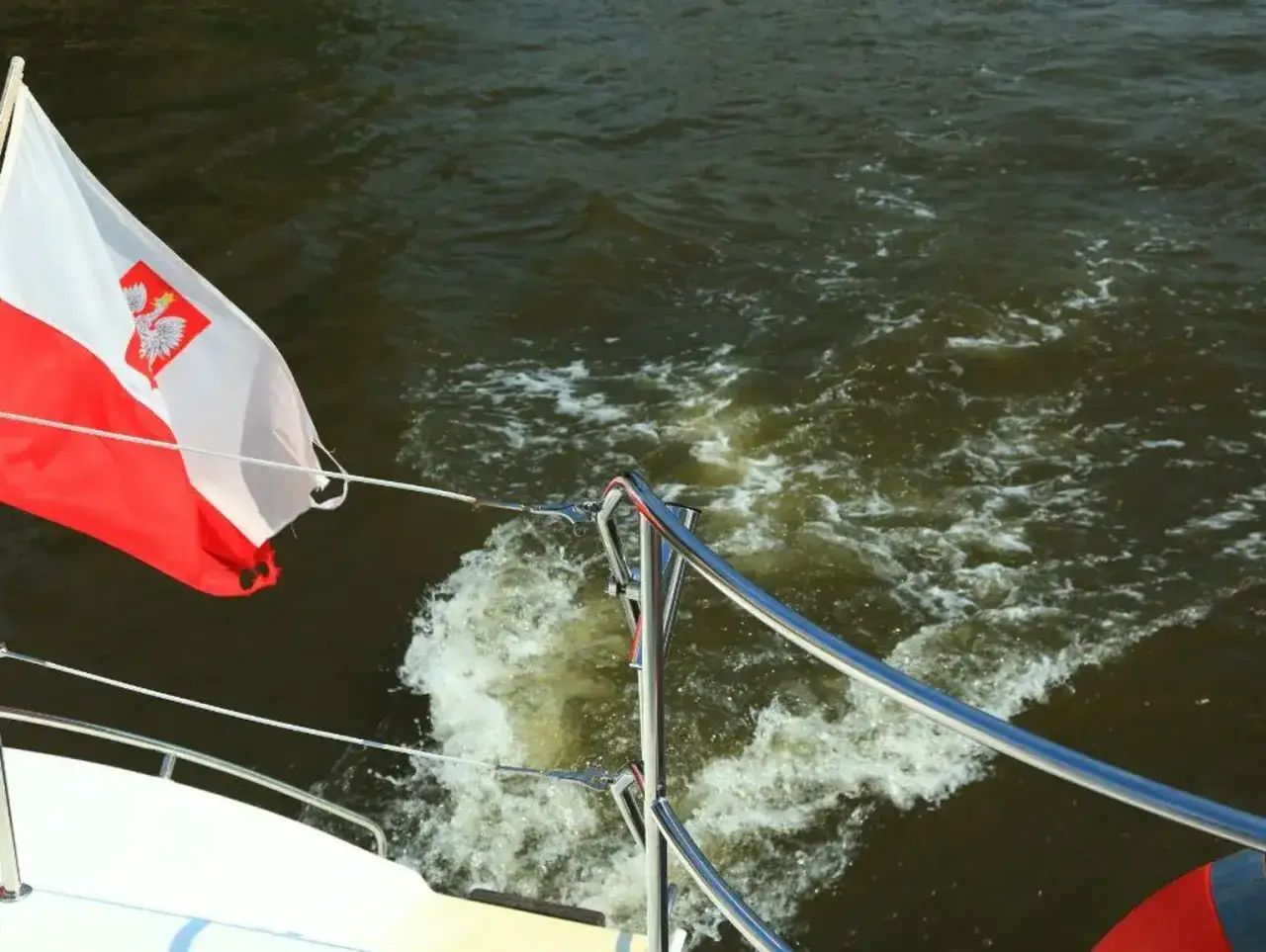 Flaga polska powiewa na wietrze, gdy łódź pruje wodę, tworząc białą smugę.