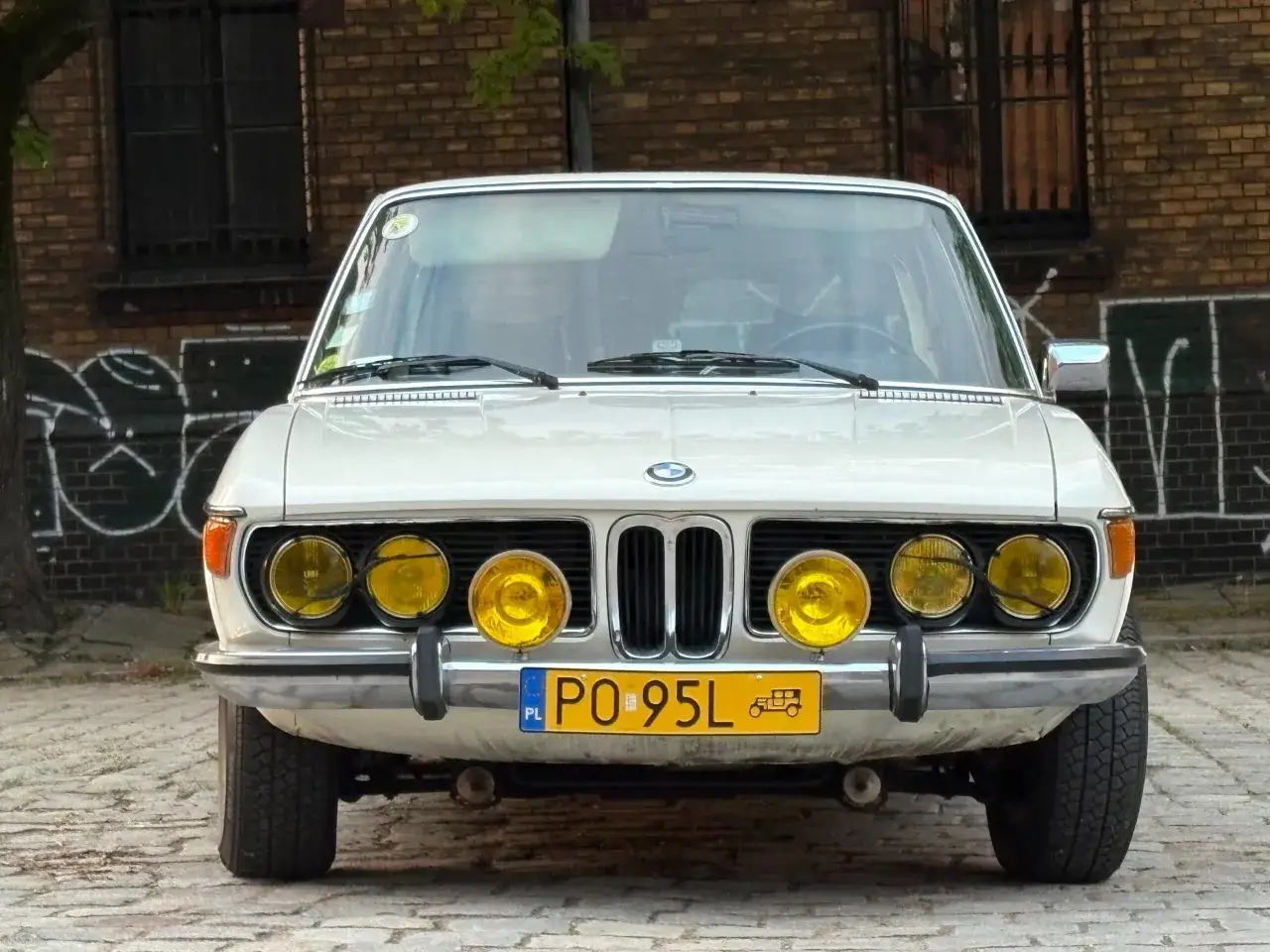 Klasyczny biały BMW E3 z czterema żółtymi światłami drogowymi i polską tablicą rejestracyjną PO 95L.
