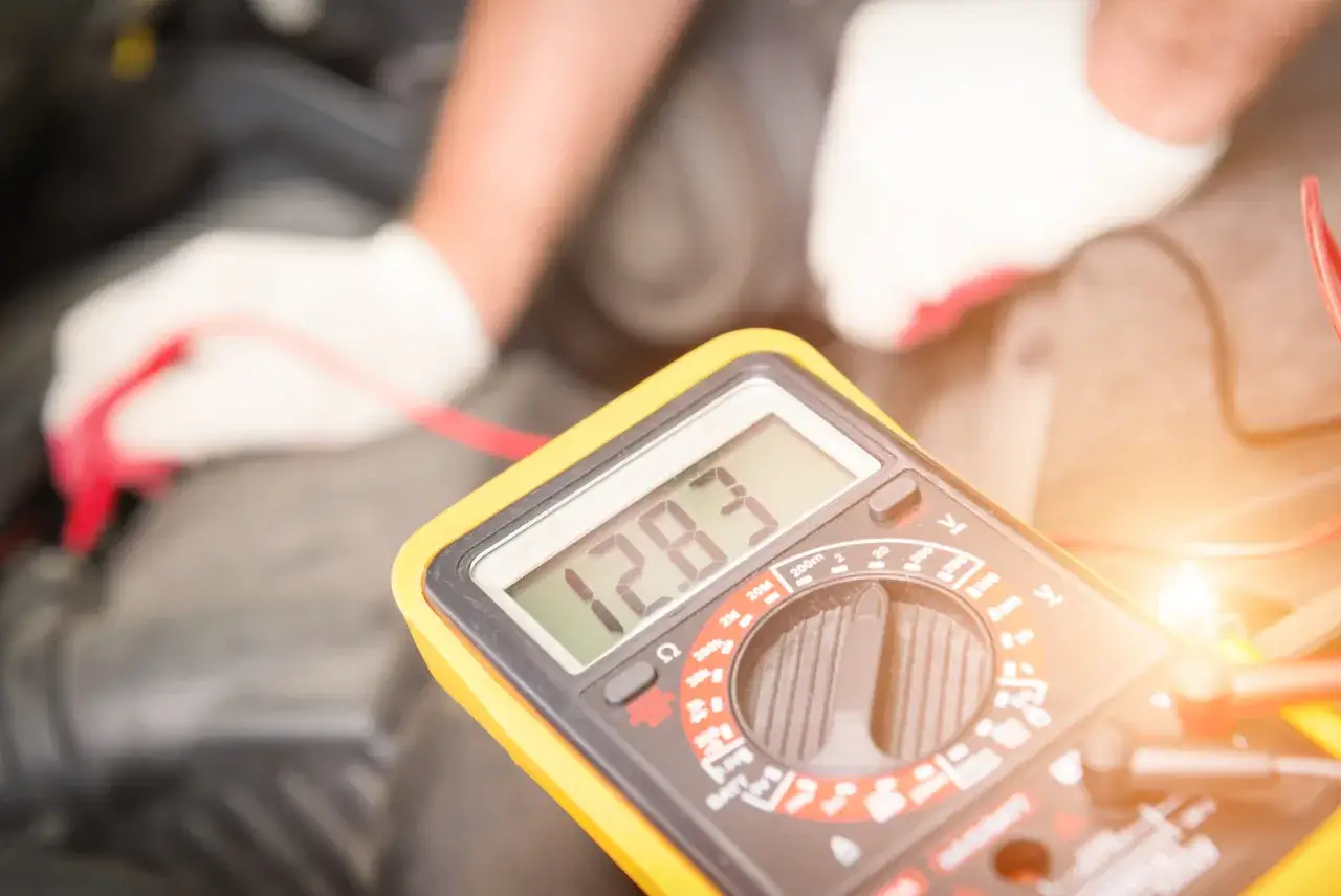 Mechanik w białych rękawiczkach mierzy napięcie akumulatora multimetrem. Wynik 12.83V sugeruje, że problem z tym, że po przekręceniu kluczyka nie świecą kontrolki, leży gdzie indziej.